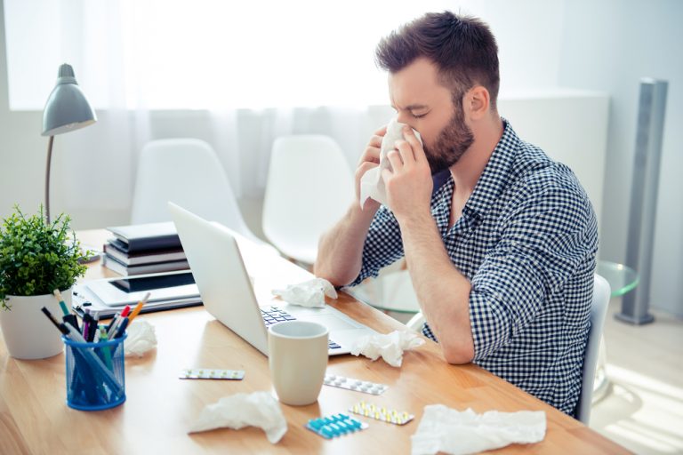Hombre estornudando sobre un escritorio mientras trabaja en casa