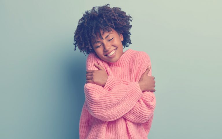 Mujer afro con un buzo rosado dándose un abrazo