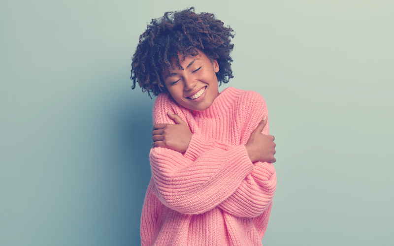 Mujer afro con un buzo rosado dándose un abrazo