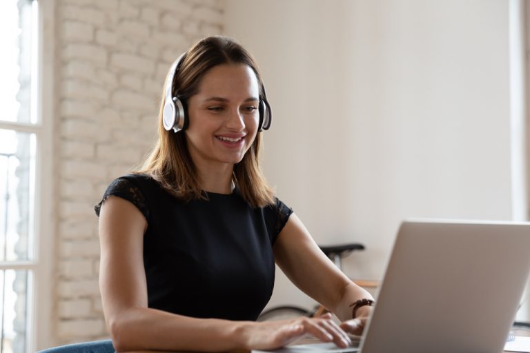 Mujer sonriente con audífonos inalámbricos trabajando escribiendo en un portátil sentado en el escritorio en el trabajo de oficina