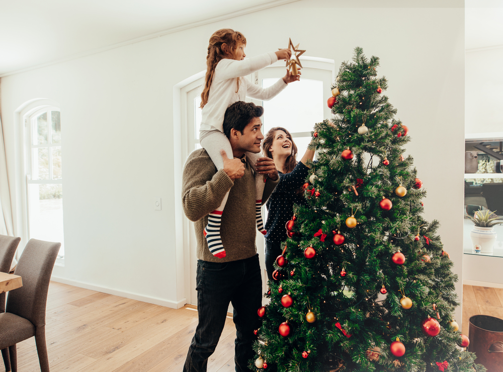 Joven con su hija en los hombros ayudándola a decorar el árbol de Navidad.