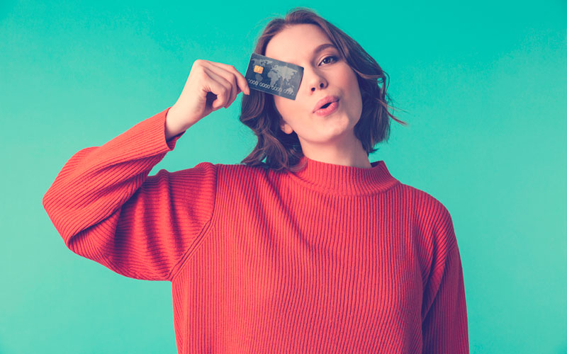 Retrato de una mujer muy joven vestida con suéter sosteniendo una tarjeta de crédito en su cara aislada sobre fondo azul