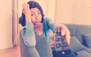 Mujer con expresión de cansancio y aburrimiento sosteniendo un control remoto en el sofá, ilustrando las consecuencias del sedentarismo.