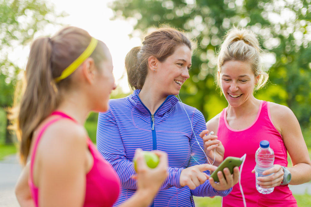 Grupo de chicas activas felices preparándose para una carrera en la naturaleza eligiendo música en un teléfono inteligente