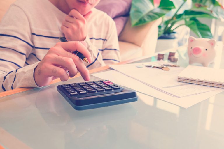 Hombre pensativo sentado haciendo cuentas en la calculadora junto con facturas, monedas y una alcancía.