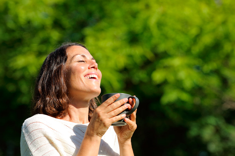 Mujer adulta satisfecha respirando aire fresco sosteniendo una taza de café al aire libre en el parque