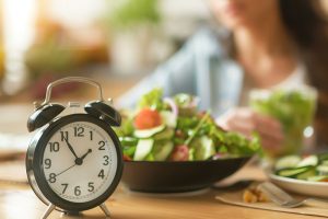 Mujer consumiendo una ensalada junto a un reloj despertador, representando los periodos de ingesta en el ayuno intermitente.