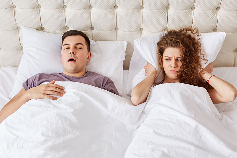 Hombre con ronquidos fuertes y signos de apnea del sueño mientras su pareja intenta dormir cubriéndose los oídos.