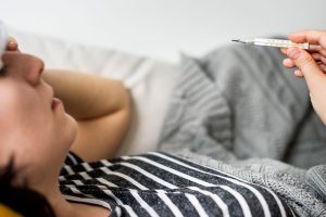 Mujer con fiebre revisando termómetro en cama, uno de los síntomas iniciales de la fiebre amarilla.