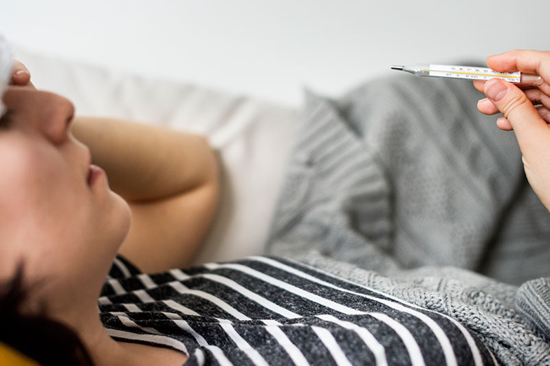 Mujer con fiebre revisando termómetro en cama, uno de los síntomas iniciales de la fiebre amarilla.