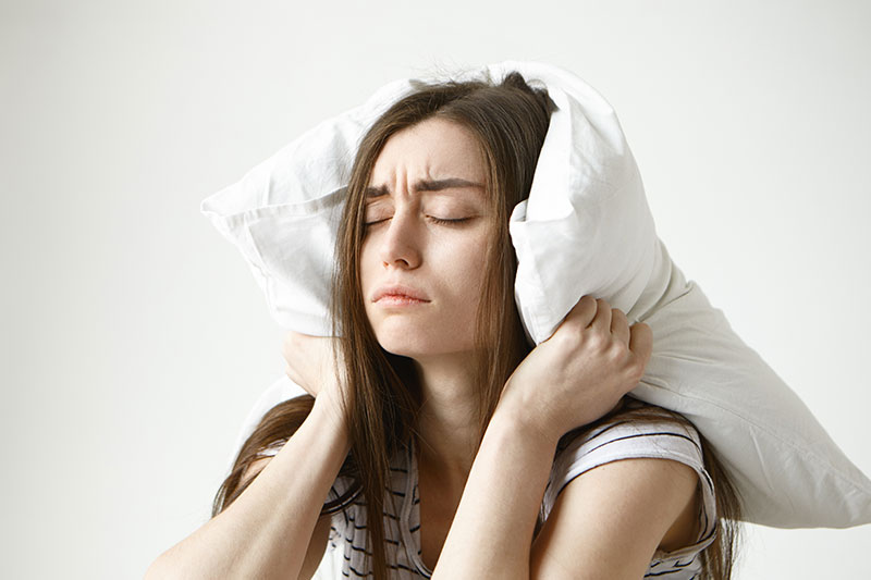 Mujer con insomnio se tapa las orejas con una almohada con expresión desesperada, ilustrando los trastornos del sueño en mujeres.