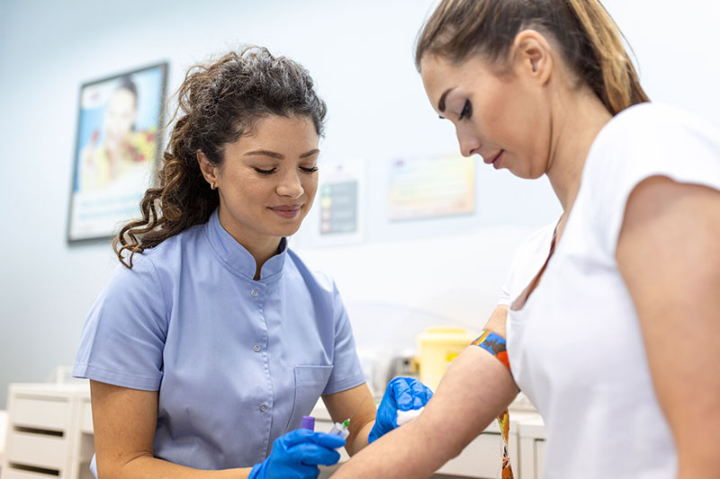 Una profesional de la salud con uniforme azul y guantes, realizando una extracción de sangre a una paciente para diagnosticar qué es la hemofilia. La escena muestra un entorno médico limpio y profesional.