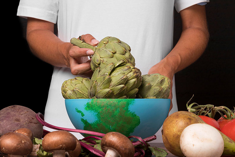 Persona sosteniendo vegetales frescos sobre un bowl con el mapa del mundo, representando la gastronomía sostenible y alimentación consciente.