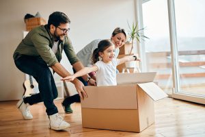 Madre y padre jugando con su hija en una caja de cartón, representando el bienestar financiero familiar y la estabilidad del hogar protegido por seguros.