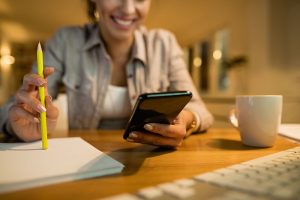 Mujer sonriendo mientras revisa su presupuesto financiero en el celular y toma notas para ahorrar y saber como usar la prima de mitad de año