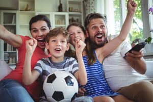 Familia feliz celebrando un gol frente al televisor sosteniendo un balón de fútbol en la sala de su casa, ilustrando ahorrar en el mundial y mantener su bienestar financiero