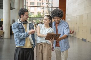 Tres profesionales jóvenes y diversos sonríen mientras colaboran revisando una tableta digital en las instalaciones modernas de una empresa que sabe cómo retener talento humano.