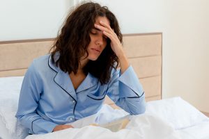 Mujer sentada en su cama con la mano en la cabeza por los síntomas premenstruales.