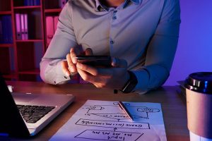 Profesional usando smartphone para integrar IA en el trabajo junto a esquema de planificación y café en escritorio.