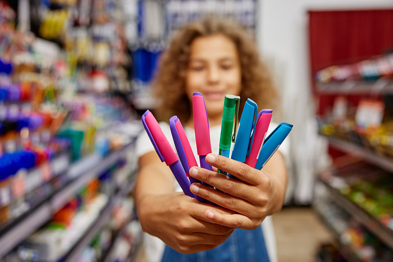 Niña sosteniendo bolígrafos de colores en una papelería durante las compras de útiles escolares.